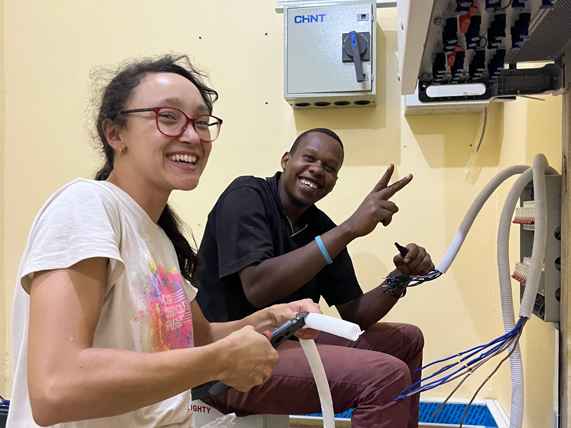 Friends smiling while working on an electrical installation, representing OMASI's energy access projects.